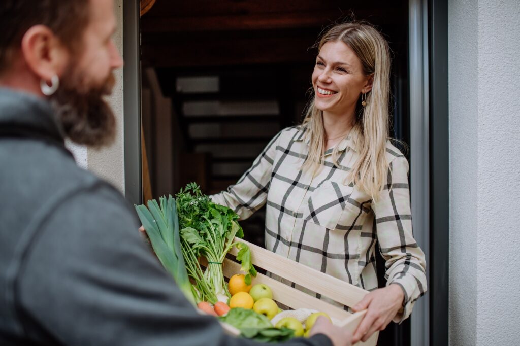 Mature man holding crate with vegetales and fruit and delivering it to woman standing at doorway.